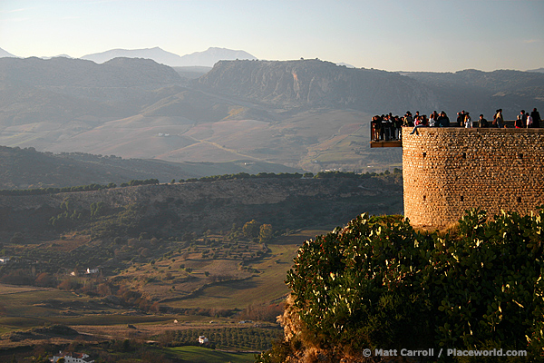 View of rugged countryside from cliff at Ronda, Spain. There is a viewing platform over edge of cliff full of people.